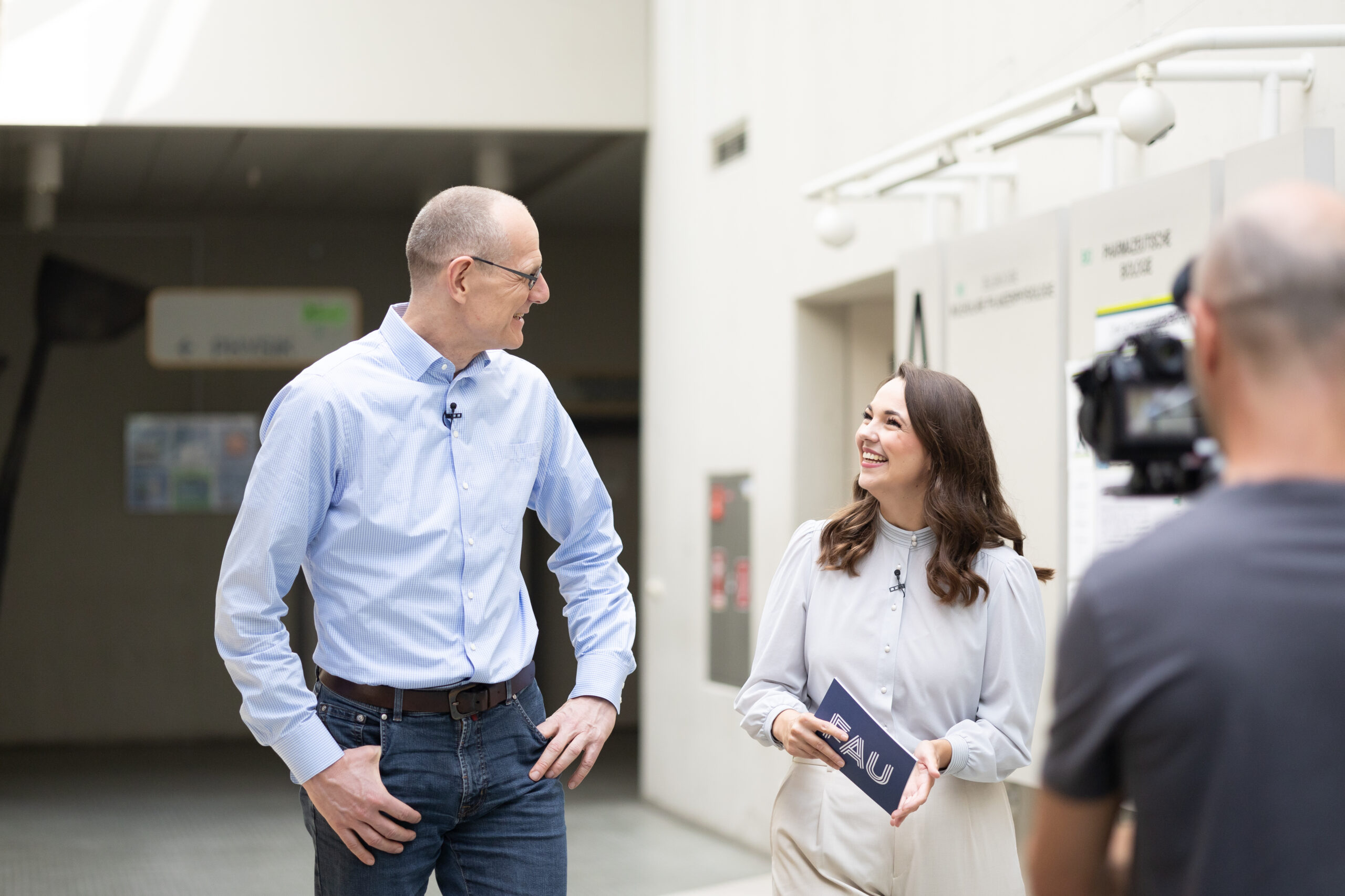 Dr. Bernd Montag an der Naturwissenschaftlichen Fakultät der FAU, zusammen mit junger Frau, die ihn interviewt. Sie befinden sich im inneren des Physikums in einem hellen Gang und laufen nebeneinander.