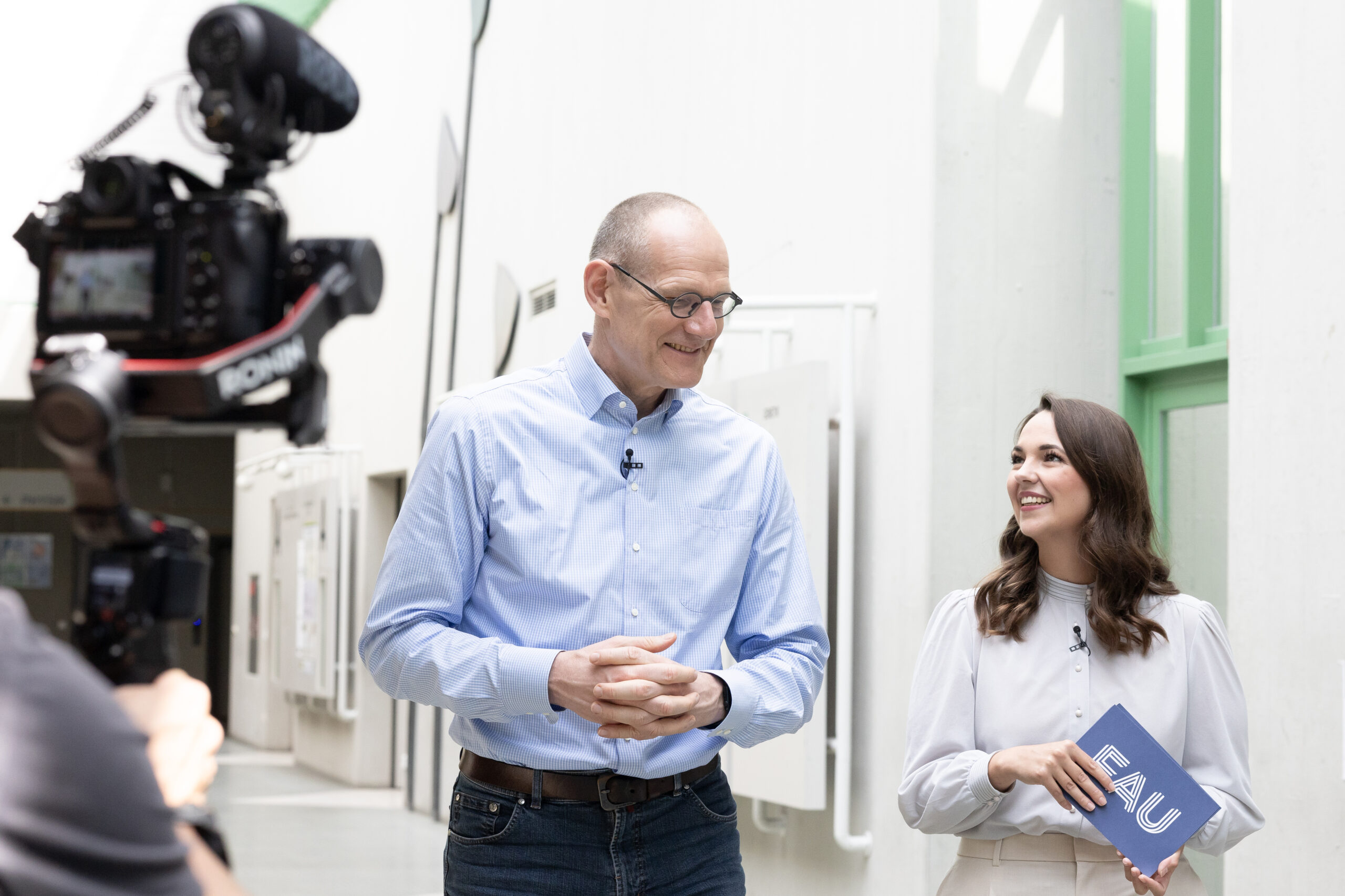Dr. Bernd Montag an der Naturwissenschaftlichen Fakultät der FAU, zusammen mit junger Frau, die ihn interviewt. Sie befinden sich im inneren des Physikums in einem hellen Gang und laufen nebeneinander.