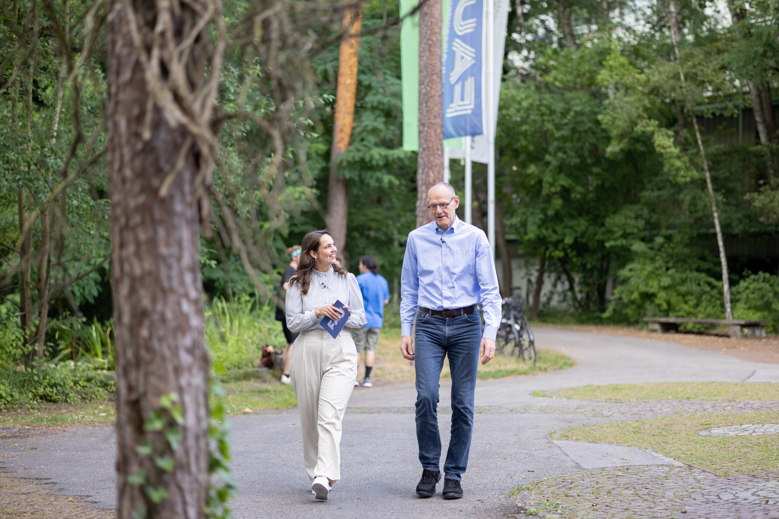 Dr. Bernd Montag an der Naturwissenschaftlichen Fakultät der FAU, zusammen mit junger Frau, die ihn interviewt. Sie befinden sich im Freien vor dem Physikum und laufen nebeneinander her.