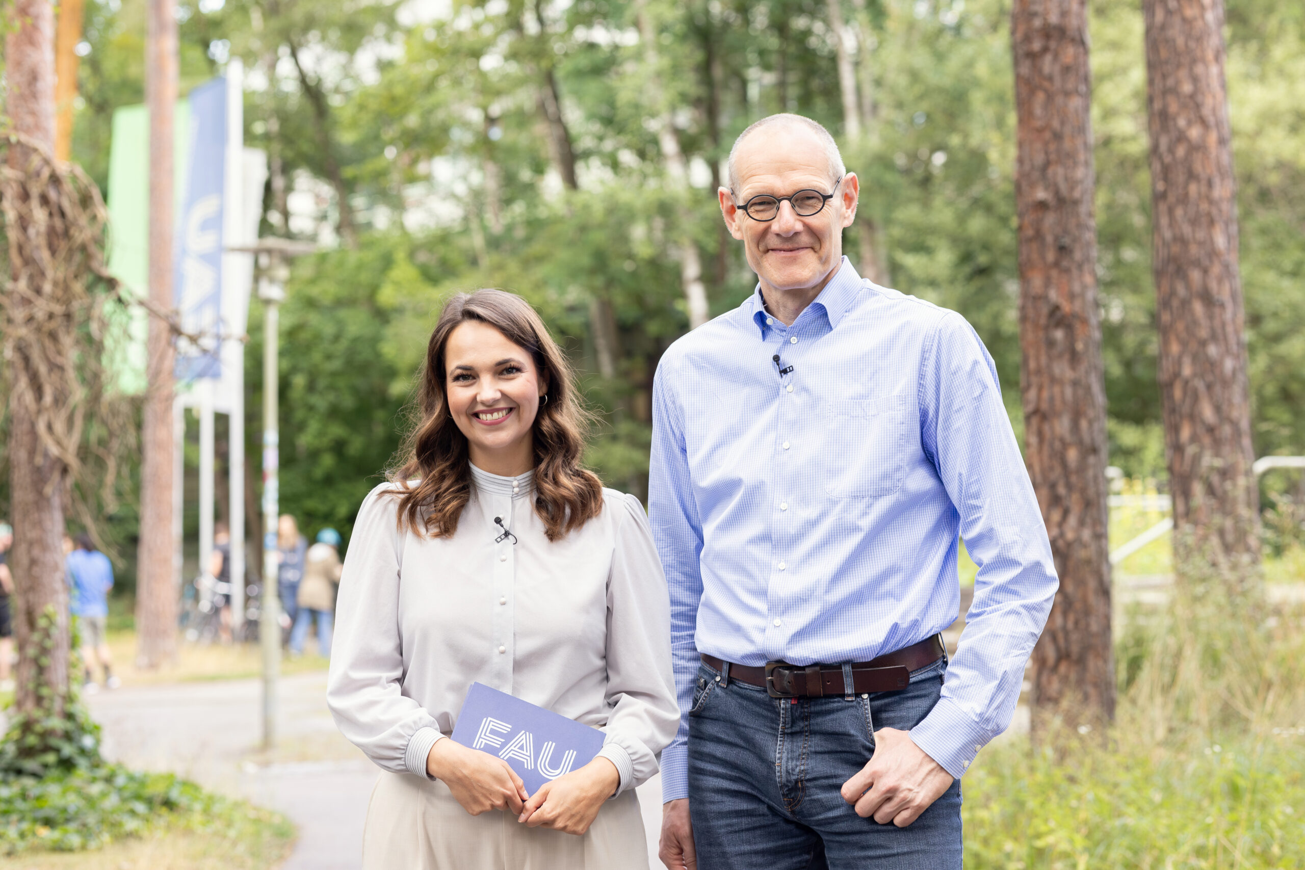 Dr. Bernd Montag an der Naturwissenschaftlichen Fakultät der FAU, zusammen mit junger Frau, die ihn interviewt. Sie befinden sich im Freien vor dem Physikum und lächeln nebeneinder stehend in die Kamera.