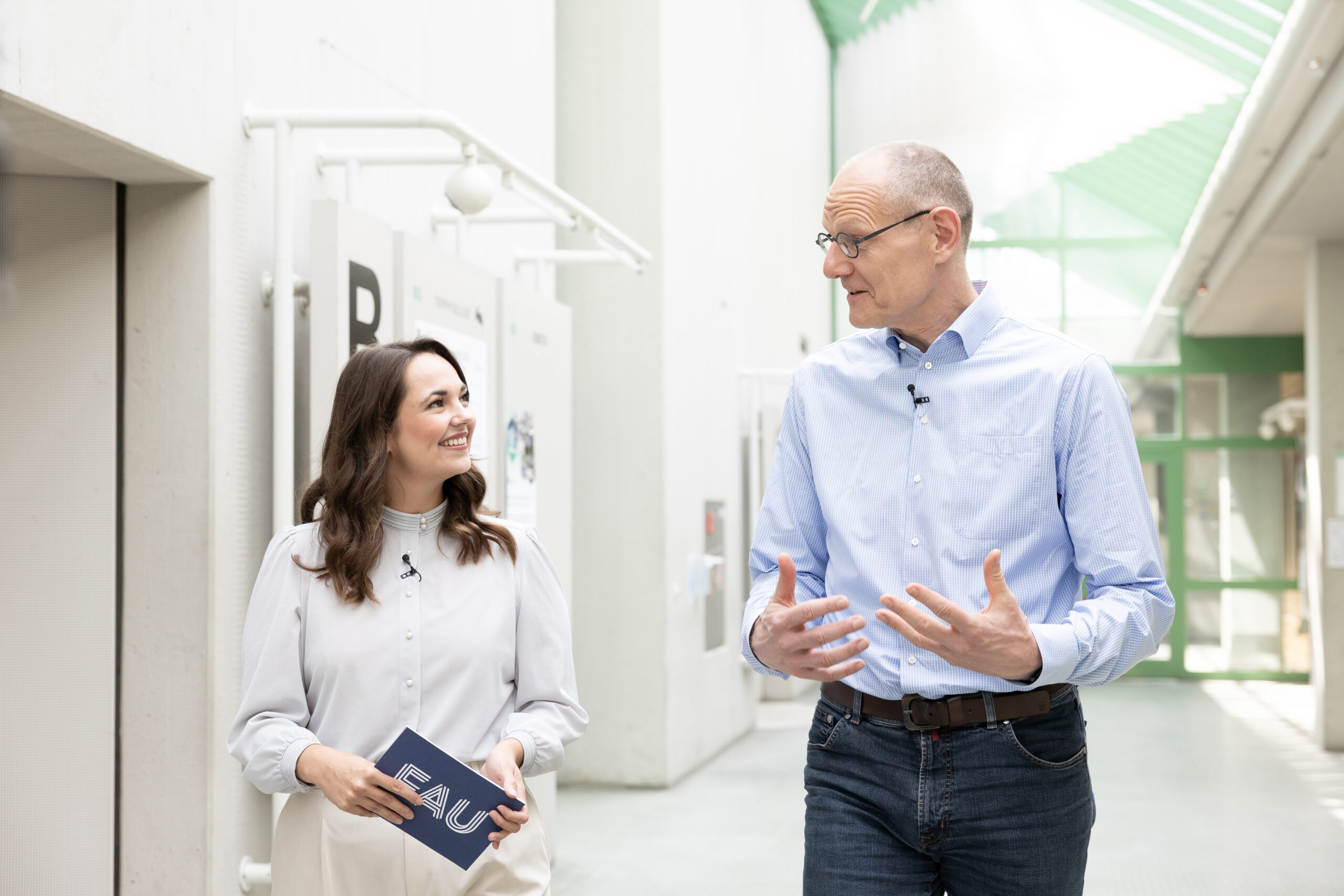 Dr. Bernd Montag an der Naturwissenschaftlichen Fakultät der FAU, zusammen mit junger Frau, die ihn interviewt. Sie befinden sich im inneren des Physikums in einem hellen Gang und laufen nebeneinander.
