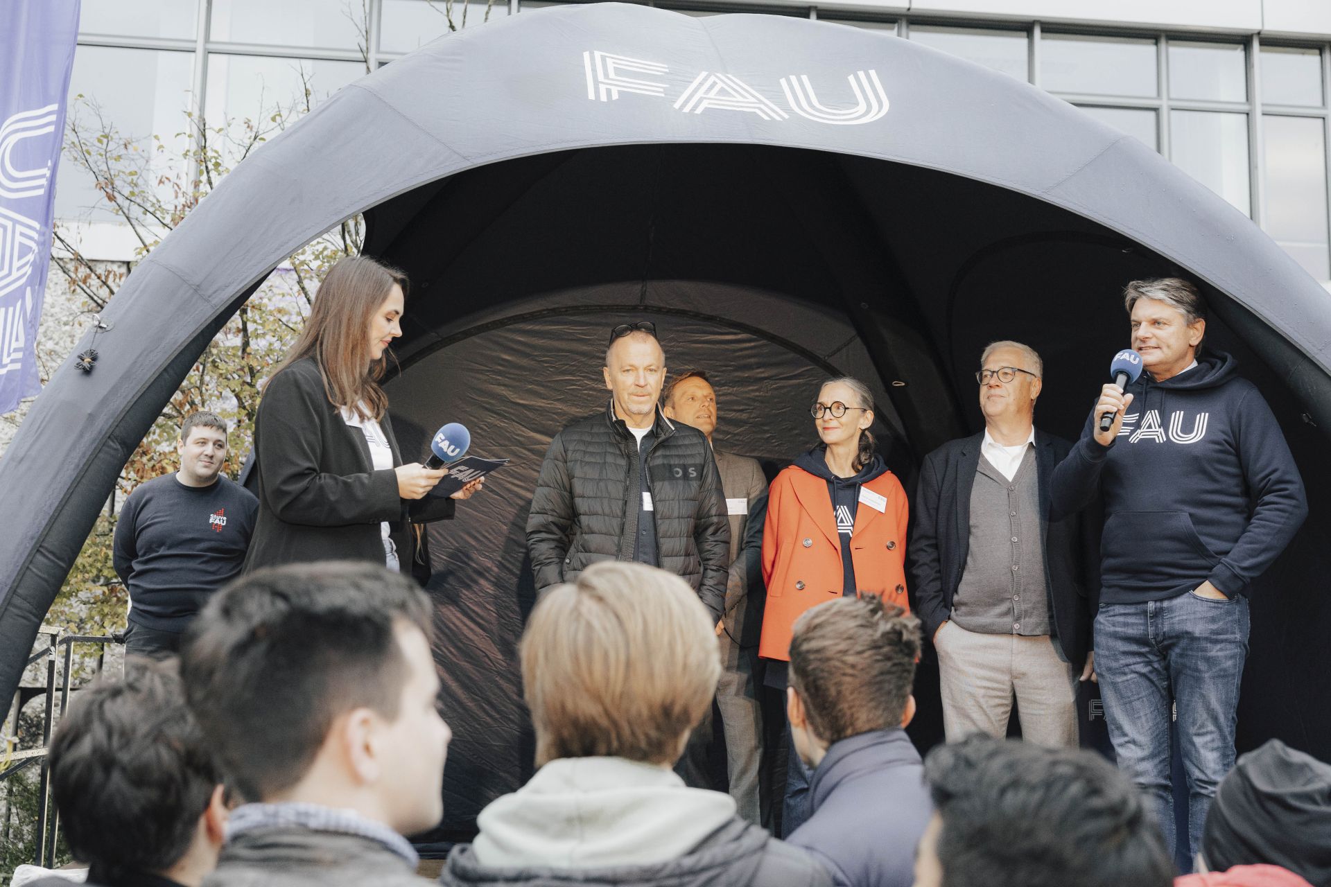 Die Universitätsleitung steht auf einer Bühne und begrüßt die Studierenden.