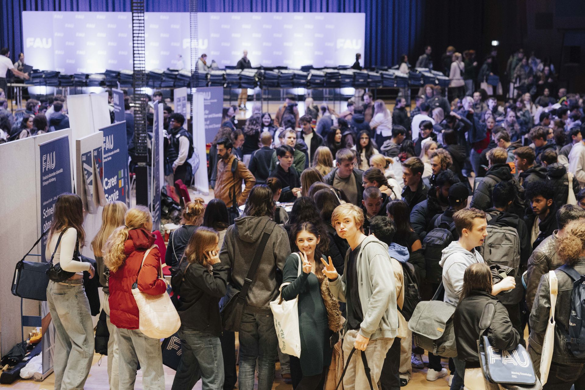 Blick in die Heinrich-Lades-Halle. Zwei Studierende grinsen in die Kamera, um sie herum viele Menschen.