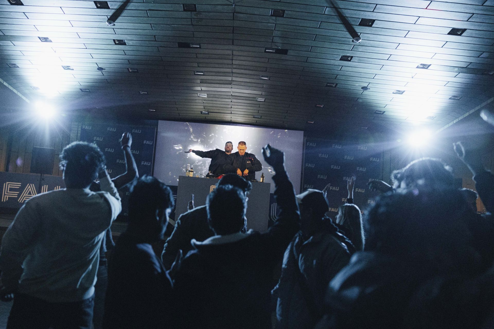 Zwei DJs stehen auf einer Bühne, Menschen davor, der Raum ist dunkel mit blauen Lichtern.