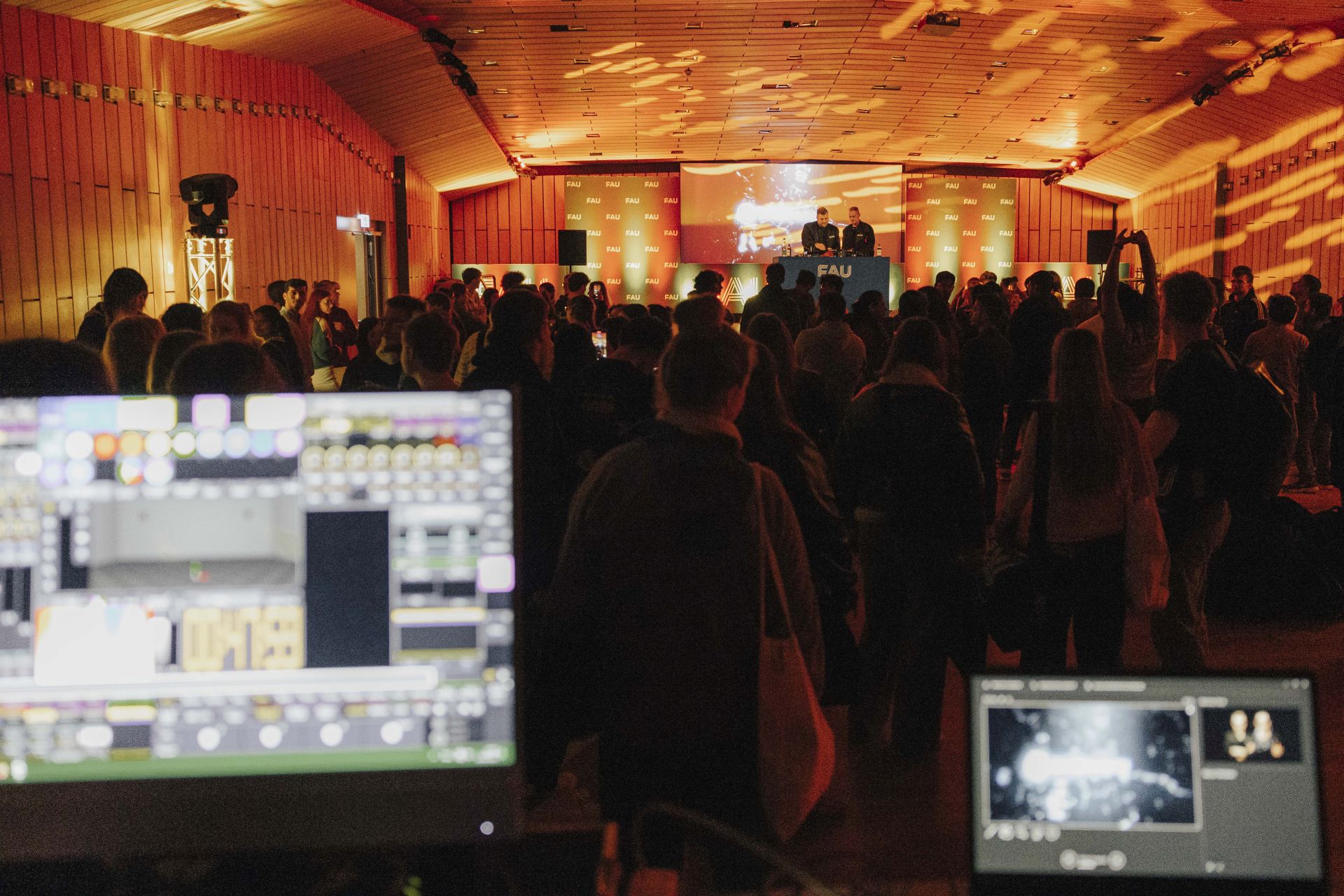 Blick auf die Bühne, wo ein DJ-Set aufgebaut ist. Der Raum ist dunkel mit orangenen Lichtern.