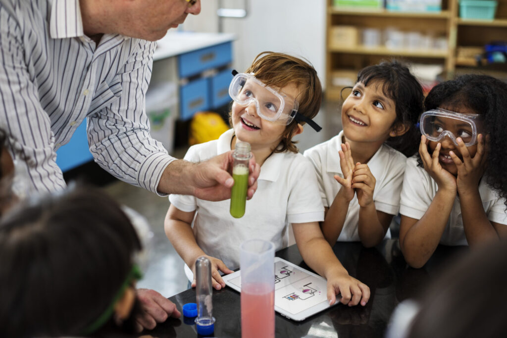 Fröhliche Kinder bei Experiment mit Flüssigkeit, die ein Forscher in einem Plastikkolben in der Hand hält. Die Kinder tragen teilweise Schutzbrillen. Kinderuni der FAU