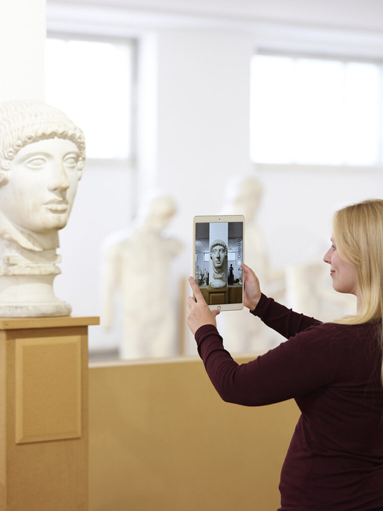 Studierende in der Antikensammlung betrachtet Gipsabguss durch Tablet.