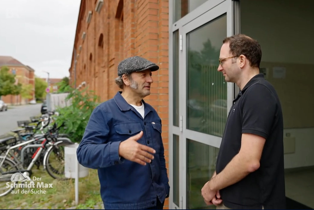 Zwei männer stehen sich gegenüber und sprechen miteinander. Schmidt Max trägt einen mütze, Dr. Reichenbach eine Brille.