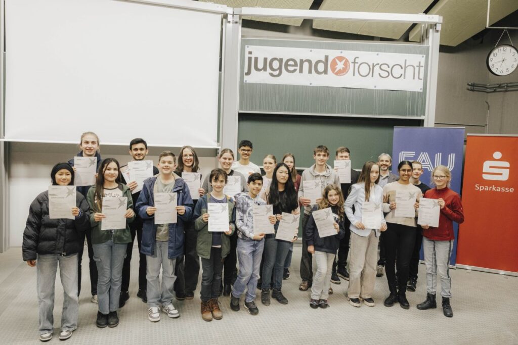 Gruppenfoto mit Kindern und Jugendlichen, die jeweils eine Urkunde in der Hand halten. Im Hintergrund eine große Tafel mit dem Schriftzug "Jugend forscht".