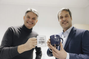 Rosshart und Hornegger halten Tassen hoch. eine tasse mit FAU -logo, die ander mit dem Aufdruck: So sieht ein richtig cooler Professor aus.