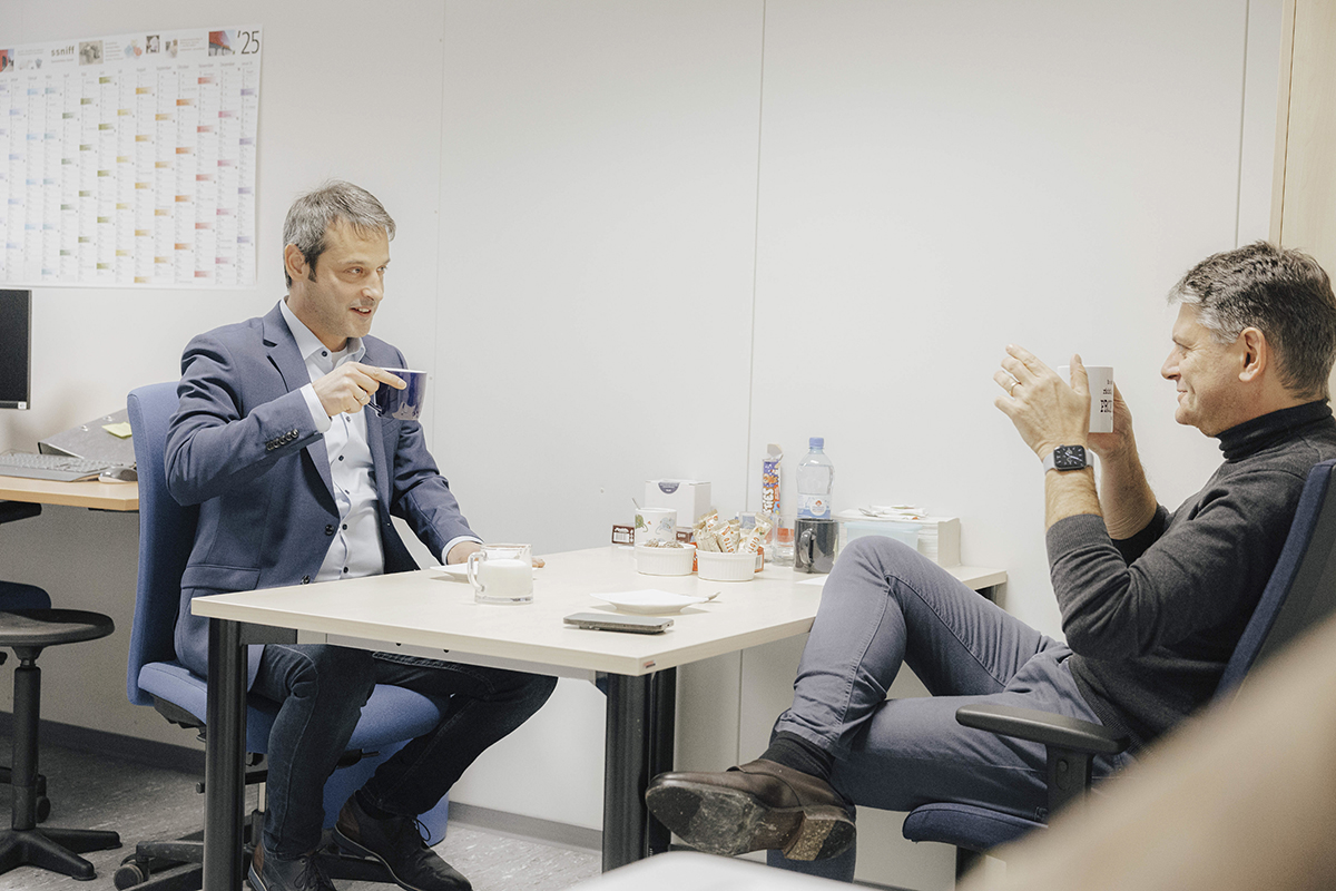 Prof Rosshart, ein Mann mit dunklen Haaren und in dunklem Anzug, im Gespräch mit FAU-Präsident Hornegger. Beide sitzen lässig an einem Tisch sich gegenüber und trinken Kaffee.