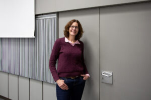 Frau Arntz, eine Frau mittleren Alters mit rotbraunen Haaren, steht an der Wand angelehnt, die Hände in den Hosentaschen.