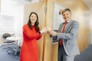 Hornegger und Handke, eine junge Frau mit dunklen Haaren und in rotem Kleid, halten gemeinsam ein Gastgeschenk von Hornegger.