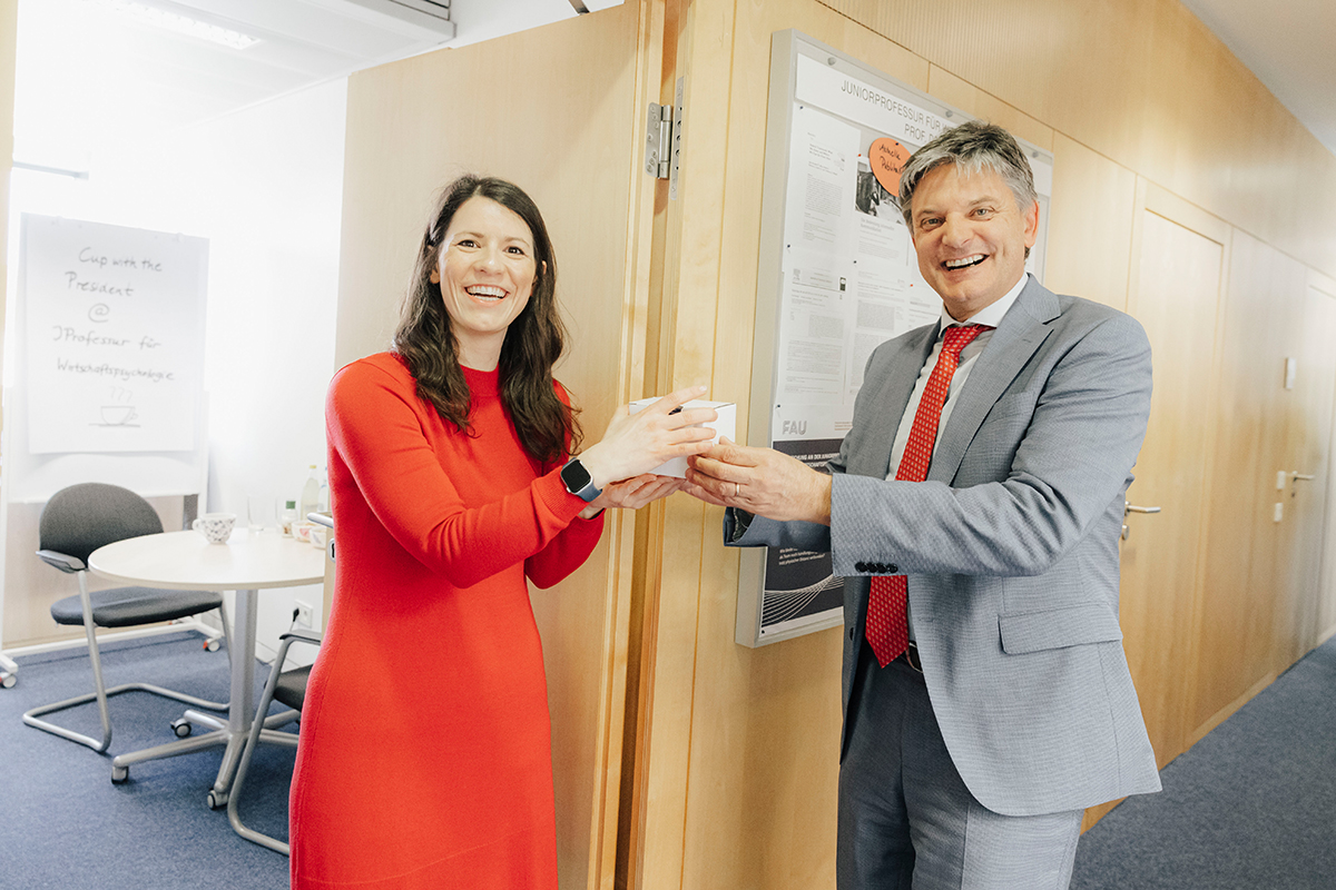 Hornegger und Handke, eine junge Frau mit dunklen Haaren und in rotem Kleid, halten gemeinsam ein Gastgeschenk von Hornegger.