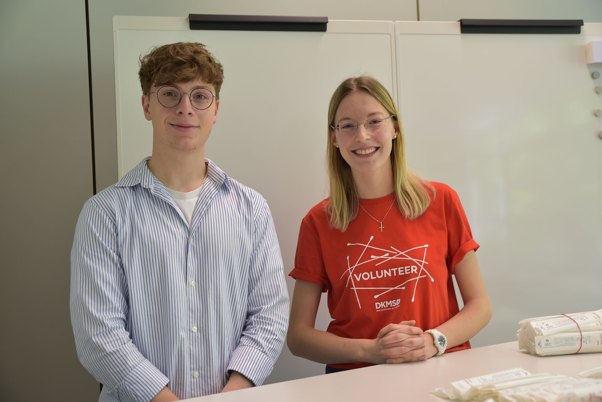 Zwei Menschen lächeln in die Kamera. Die Frau rechts trägt ein rotes DKMS Shirt.