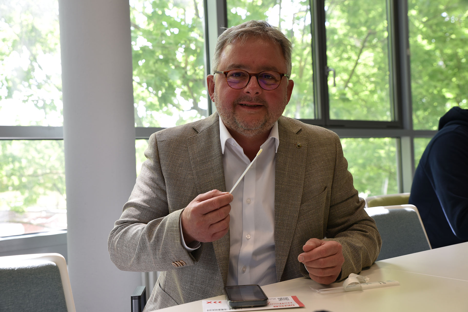 Ein Mann sitzt an einem Tisch und hält ein Wattestäbchen in die Kamera.