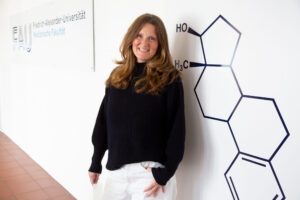 Prof. Sandforth, eine Frau mittleren Alters mit rot-braunem Haar, steht an eine Wand gelehnt da und lächelt in die Kamera.