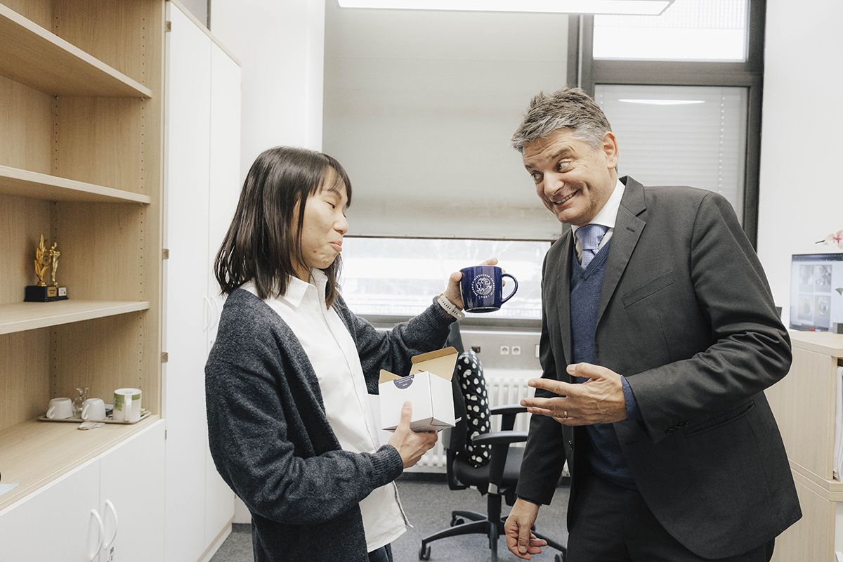 Ng und Hornegger stehen nebeneinander. Ng hält eine Tasse in der Hand und wirkt beeindruckt, Hornegger schaut sie gespannt an.