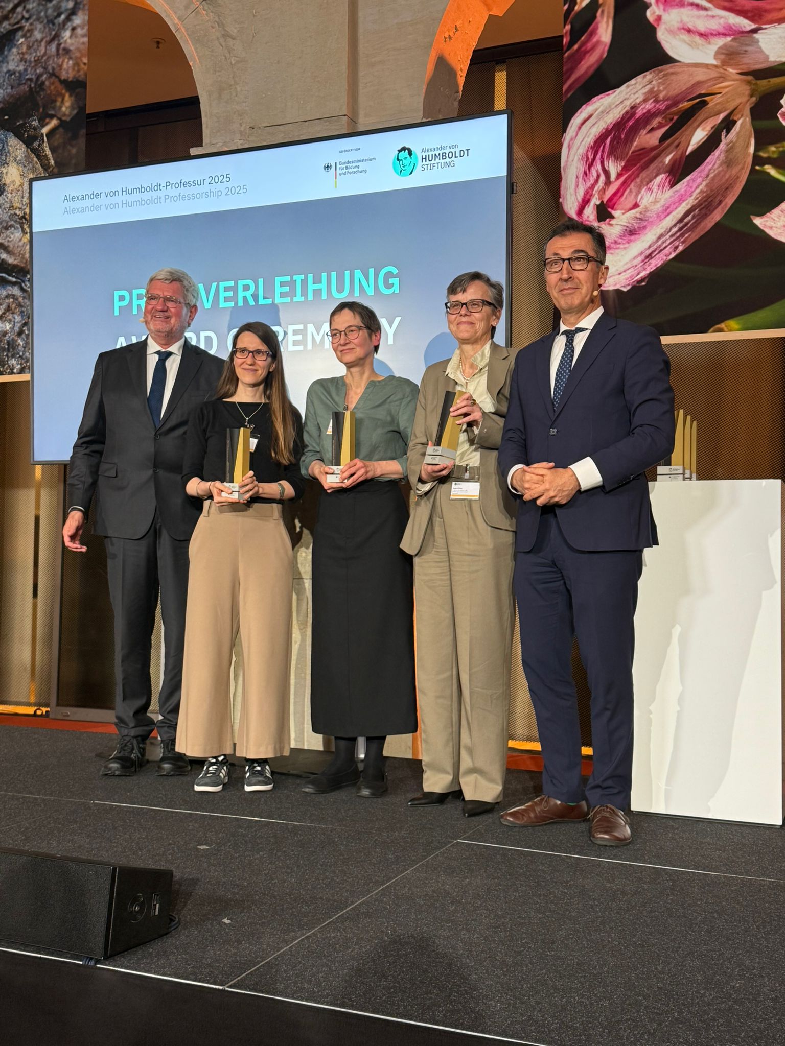 Der Präsident der Alexander von Humboldt-Stiftung Robert Schlögl steht auf einer Bühne neben den Preisträgerinnen Sandra Wachter, Eva Pils und Ingrid Piller. Die Preisträgerinnen halten jeweils eine Trophäe in den Händen. Cem Özdemir steht auf der rechten Seite. Im Hintergrund ein großer Bildschirm.
