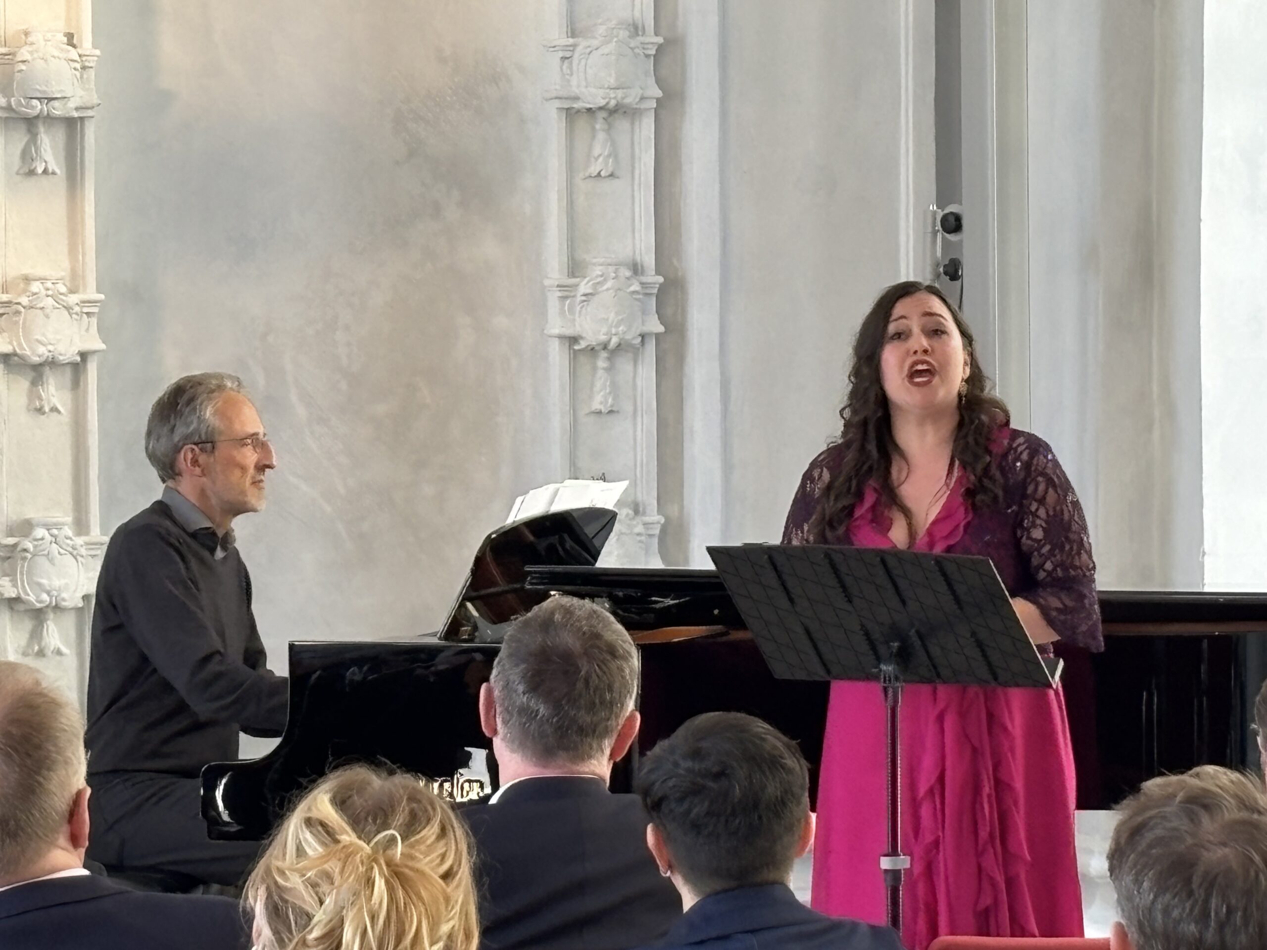 Klek, ein älterer Mann mit grau melierten Haaren am Flügel, Eismont, eine kräftige Frau im pinken Kleid, singt.