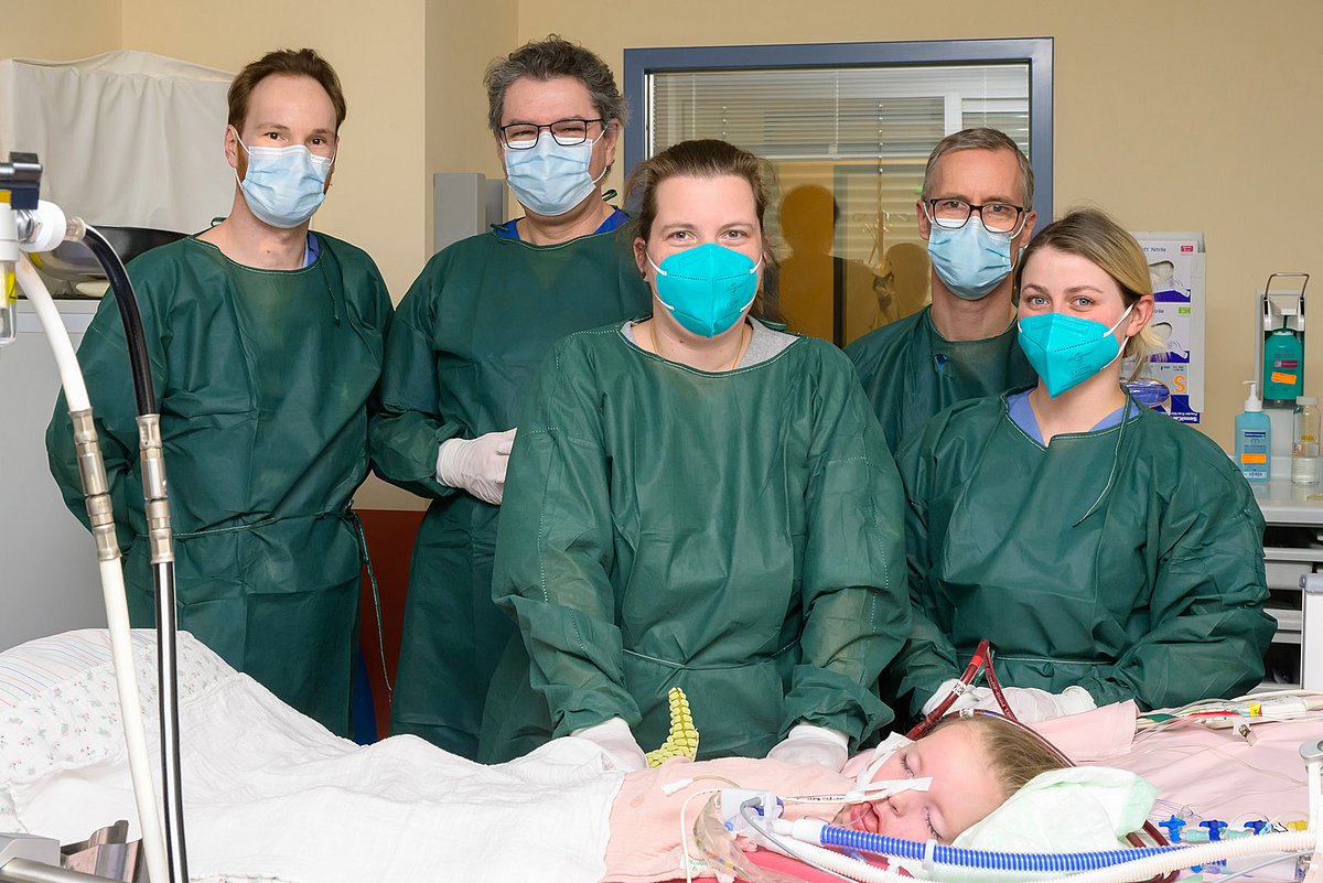 Das Ärzt:innen-Team steht mit Hannahs Mutter vor dem bett der kleinen Hannah, die immer noch intubiert ist.