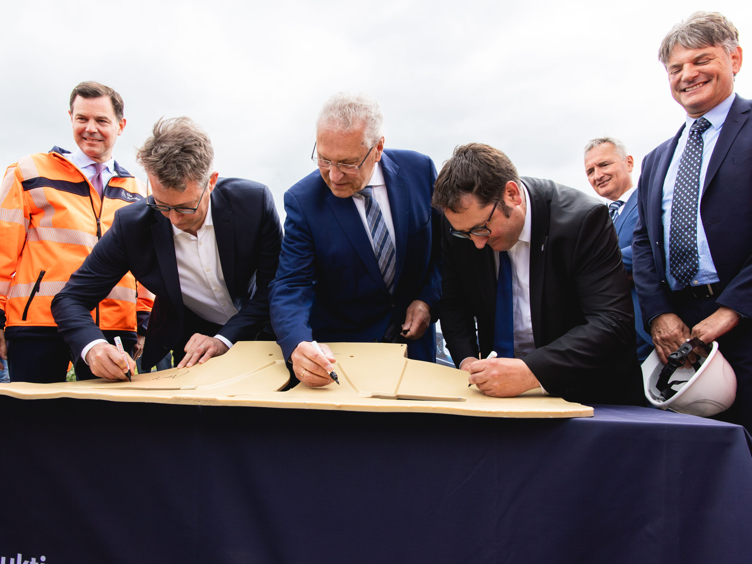 Fünf Männer in Anzügen unterschreiben auf einer symbolen Induktionsspule.