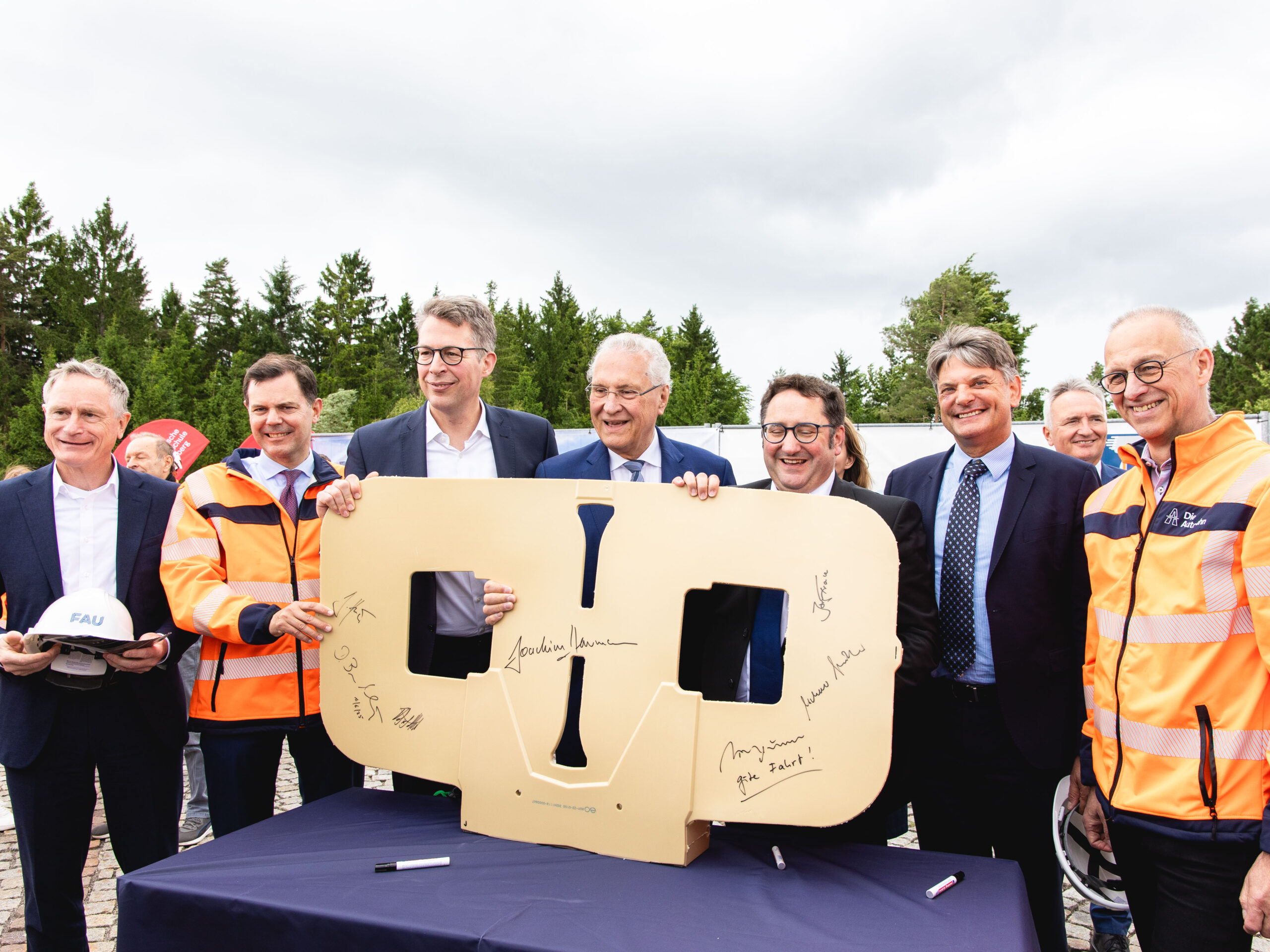 Sechs Männer in Anzügen und Warnwesten posieren mit einer symbolischen Induktinsspule