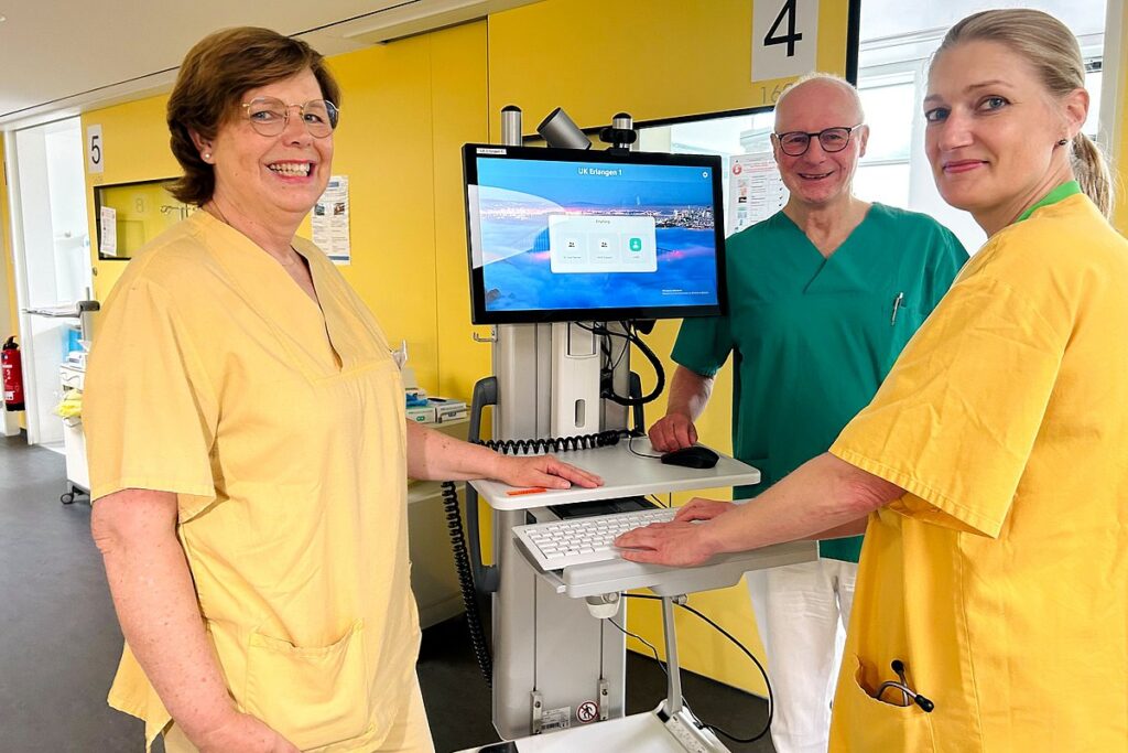 1 Arzt in grünem Kittel und 2 Ärztinnen in gelben Kitteln stehen vor einem Monitor und schauen lachend in die Kamera. 