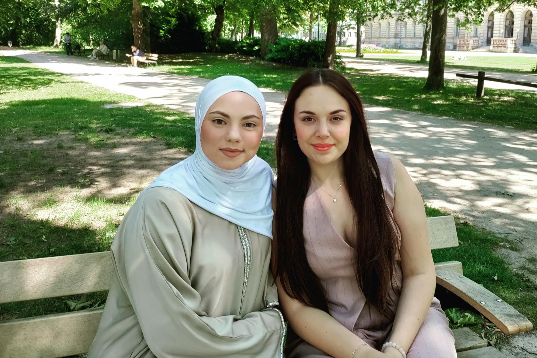 Zwei junge Frauen sitzen auf einer Parkbank im Schlossgarten und lächeln in die Kamera.