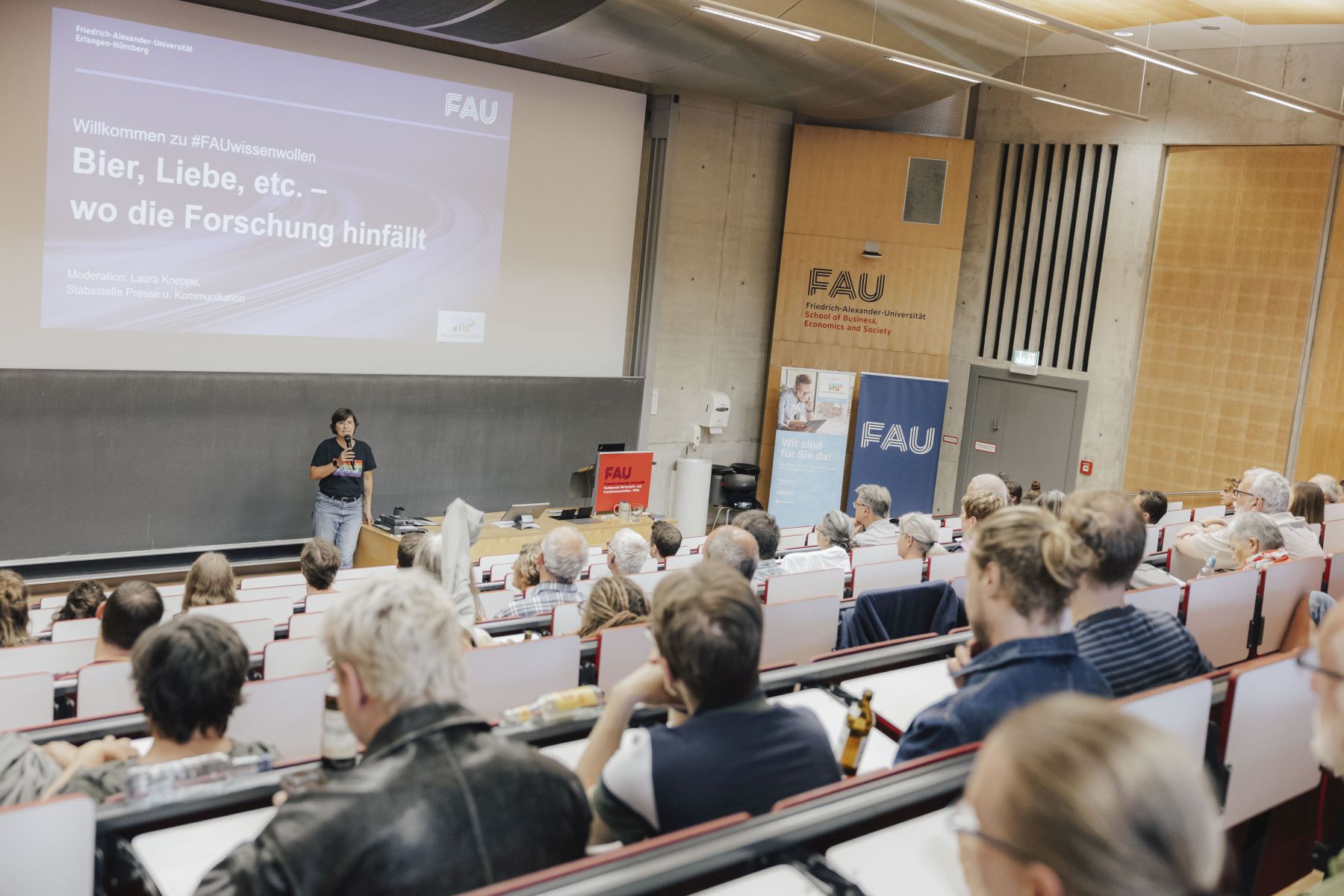 Science-Slam in FAU-Hörsaal. Präsentation im Hintergrund zeigt Folie mit Veranstaltungstitel #FAUwissenwollen. Bier, Liebe, etc. - wo die Forschung hinfällt. Vorn begrüßt die Organisatorin Laura Kneppe mit Mikrofon. Es sitzen Menschen in den Stuhlreihen.