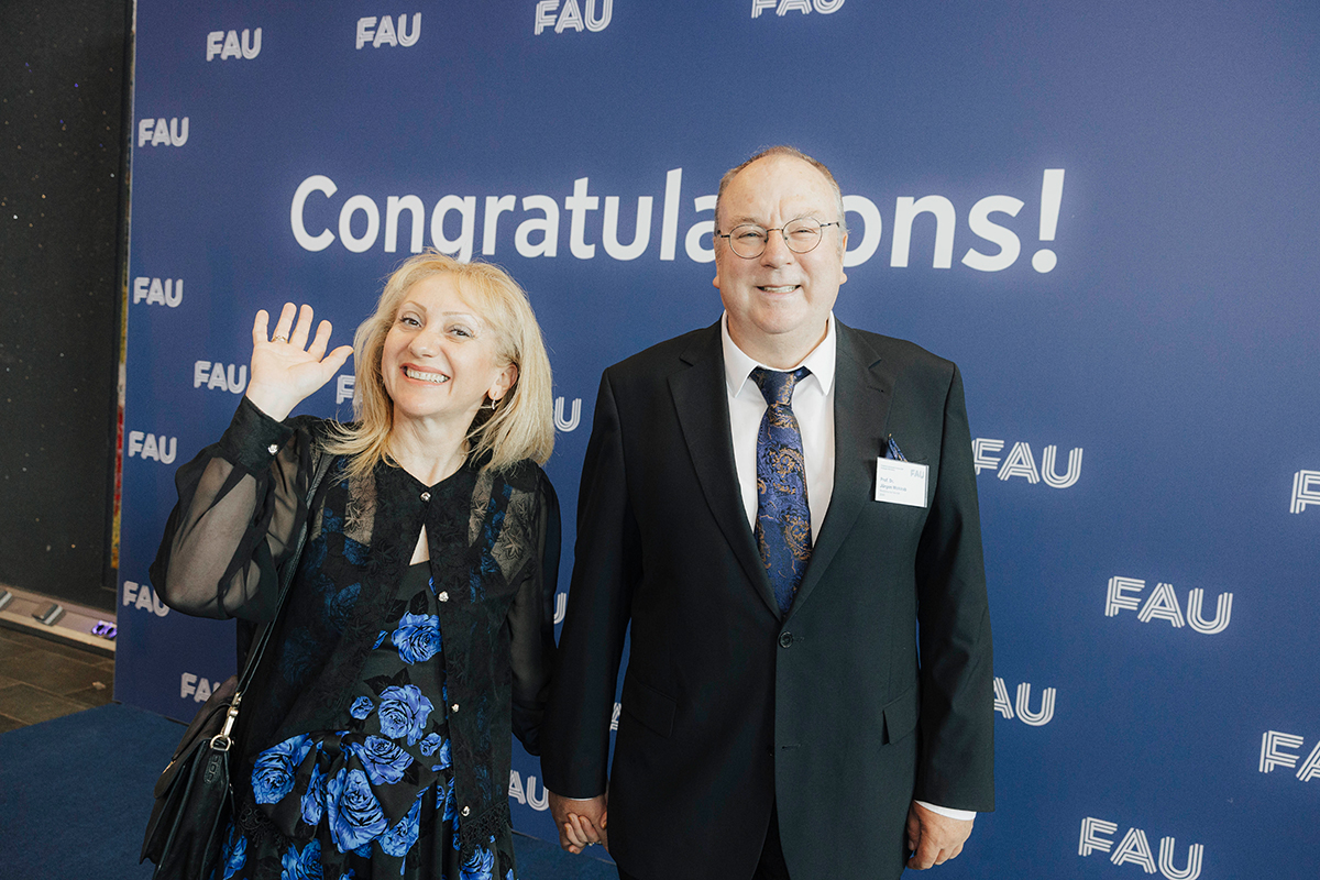 Ein Paar in Abendkleidung grüßt vor der Fotowand in die Kamera.