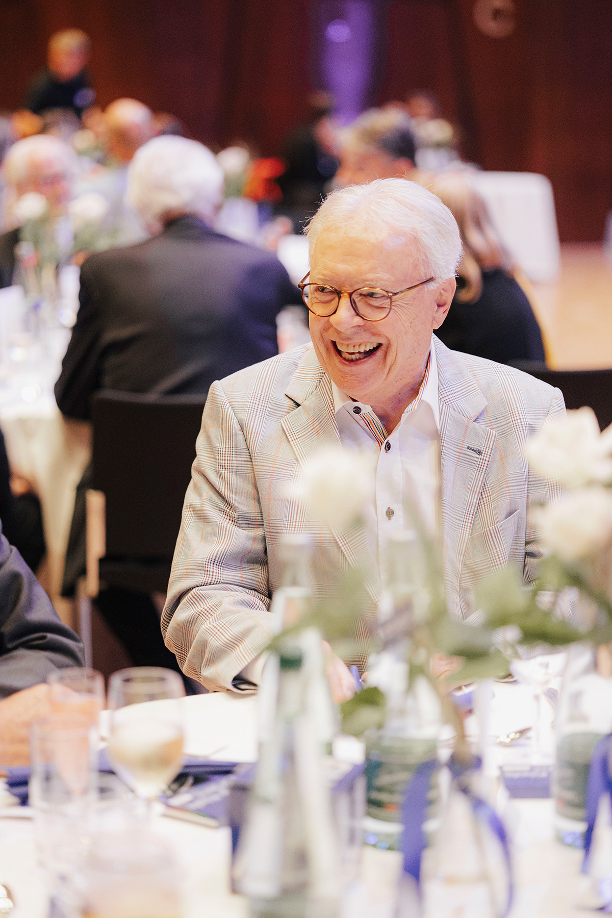 Ein Mann sitzt im beigen Sakko am Tisch und lacht.
