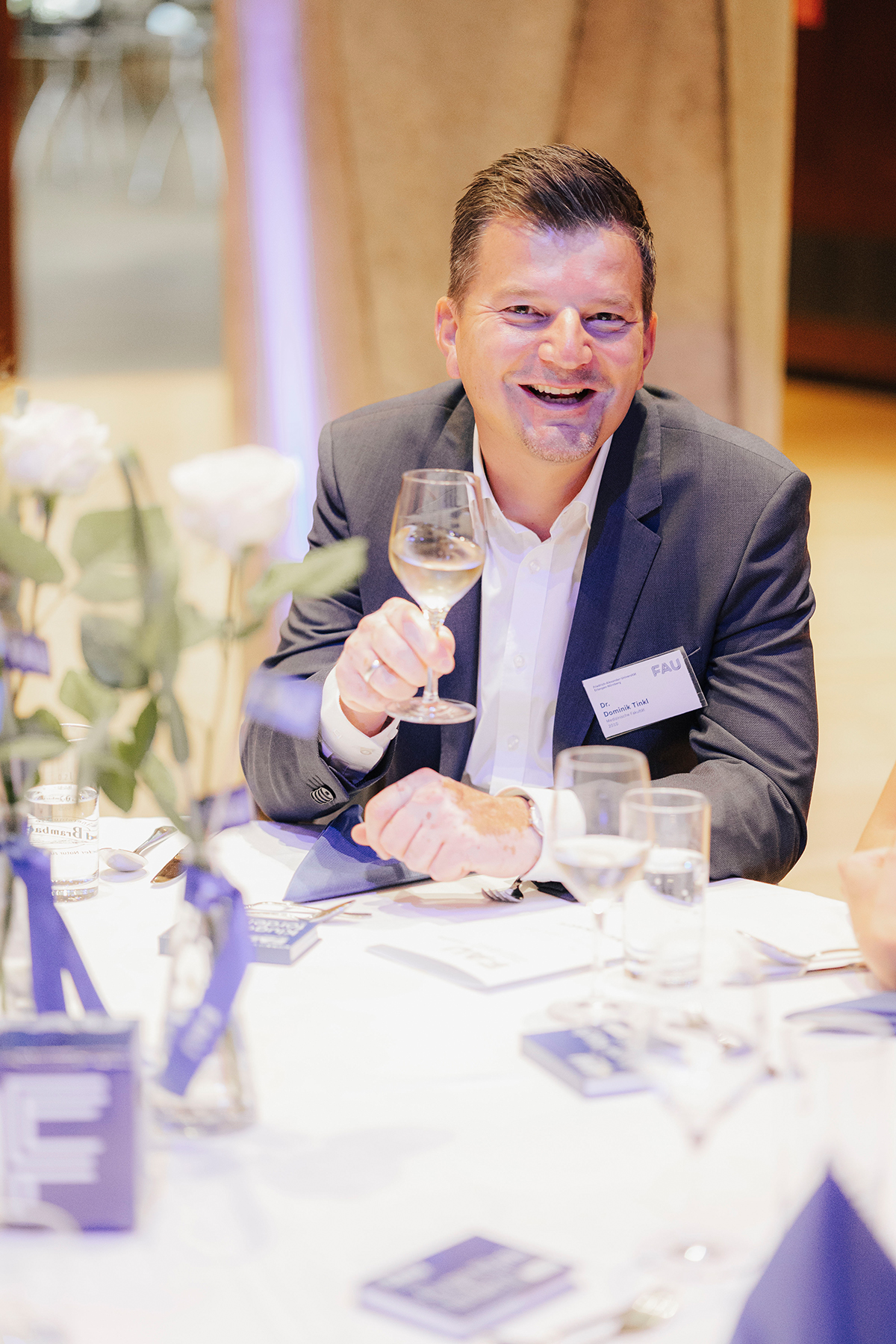 Ein Mann im grauen Sakko sitzt am Tisch und grüßt mit einem Glas