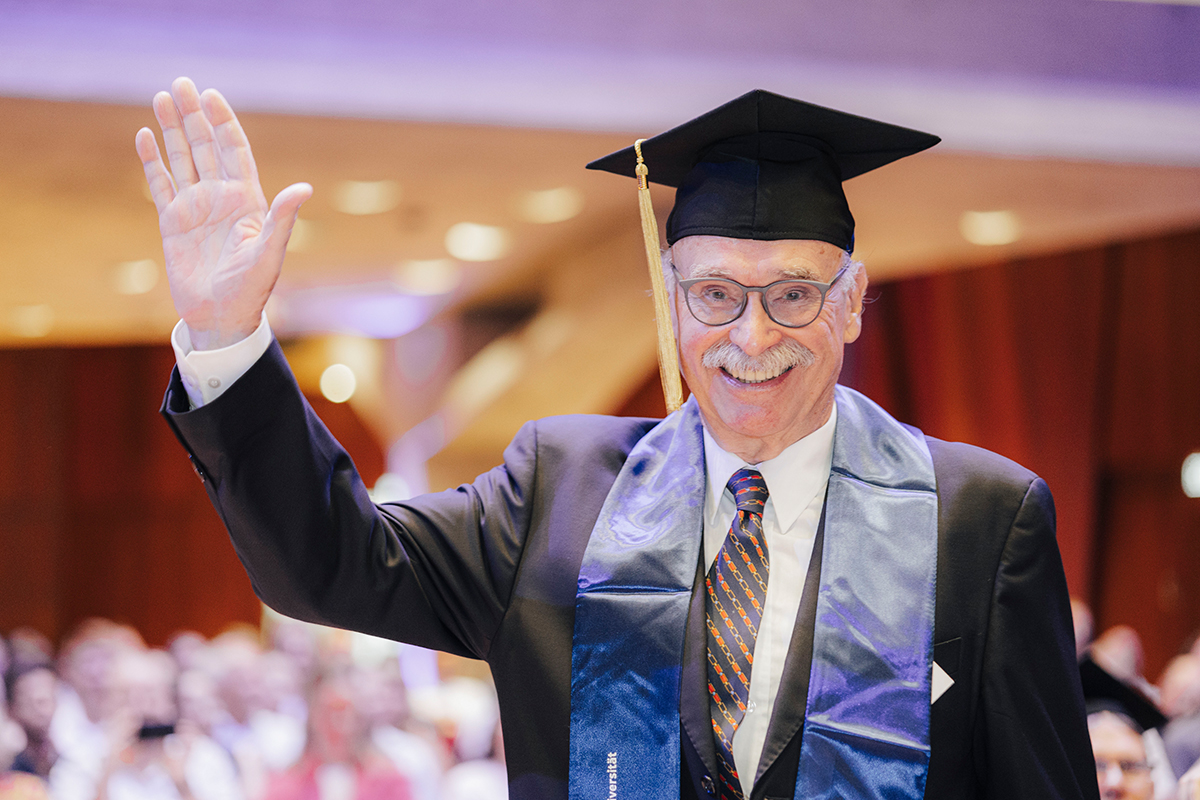 Ein älterer Jubilar hebt grüßend und lächelnd die Hand, während er feierlich Hut und Band trägt.