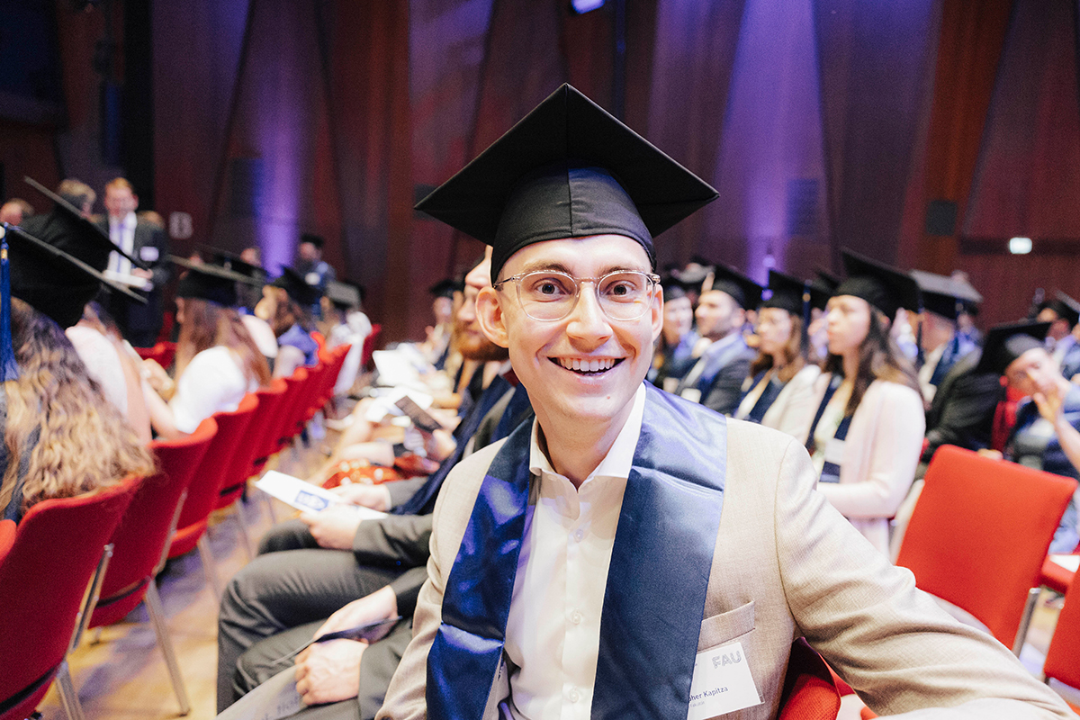 Ein junger Promovierter sitzt im Publikum und blickt lächelnd in die Kamera.