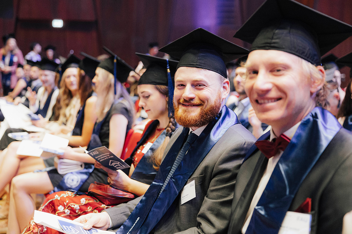 Blick in die Reihen der Promovierten, zwei Männer im Vordergrund schauen in die Kamera.