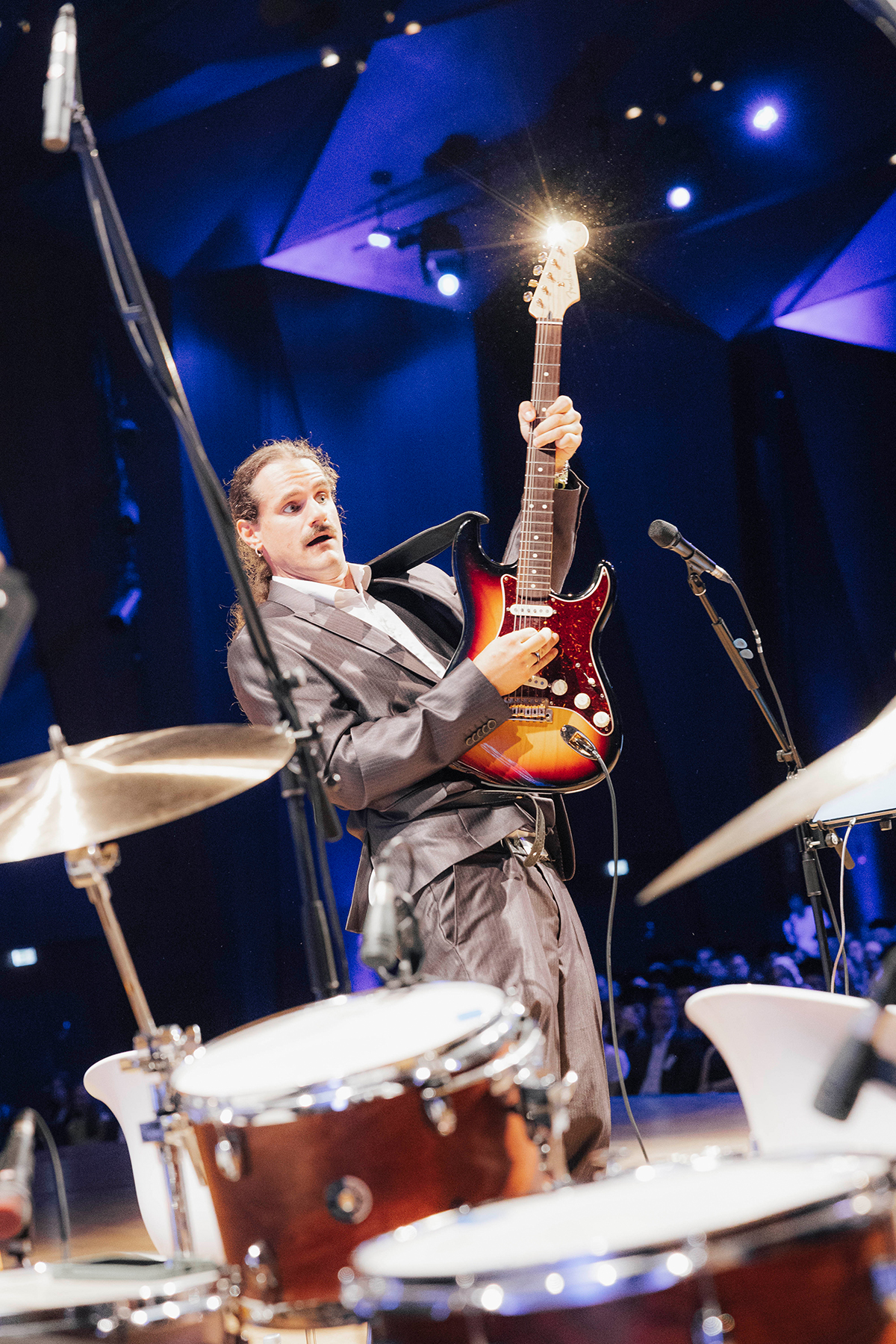 Zwischen den Drums hindurch ist der Bassist beim Spielen zu sehen.