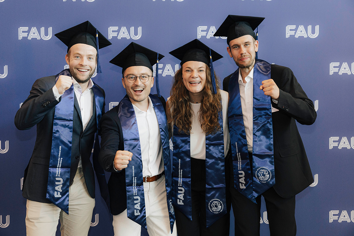 4 Jubilar*innen posieren vor der Fotowand, lächelnd, mit Band um die Schultern und Hut.