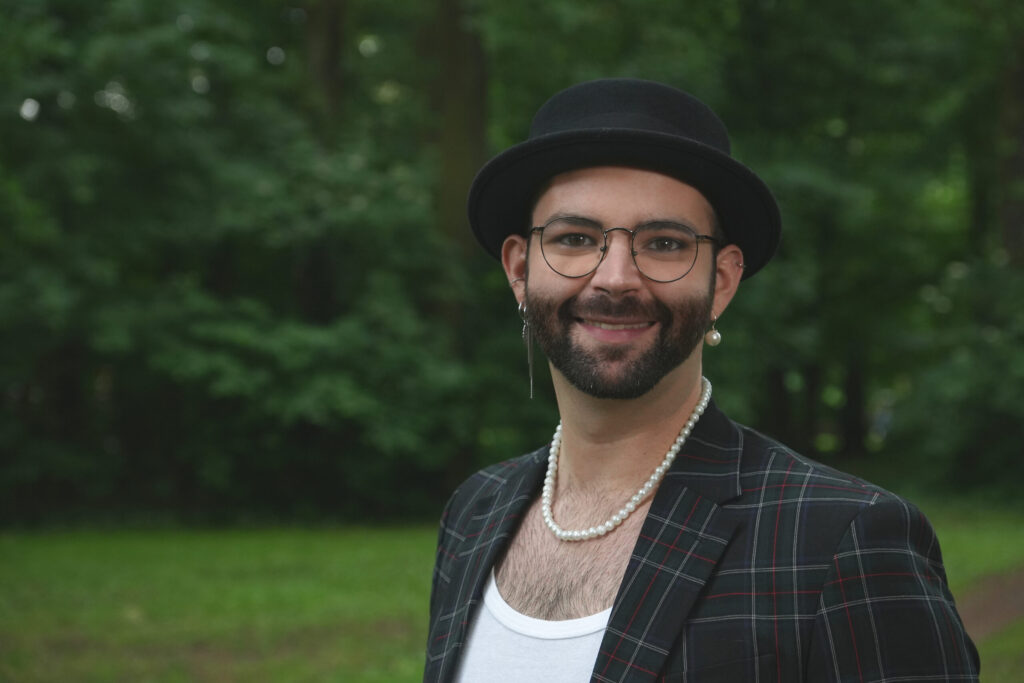 Ein dunkelhaariger, schlanker Mann zwischen 30 und 40 Jahren, mit Bart, schwarzer Brille, schwarzem Hut, Perlenkette, auffälligen Ohrringen und einen dunklen, karierten Jackett, steht auf einer grünen Wiese, man sieht den Oberkörper, im Hintergrund eine grüne Hecke.