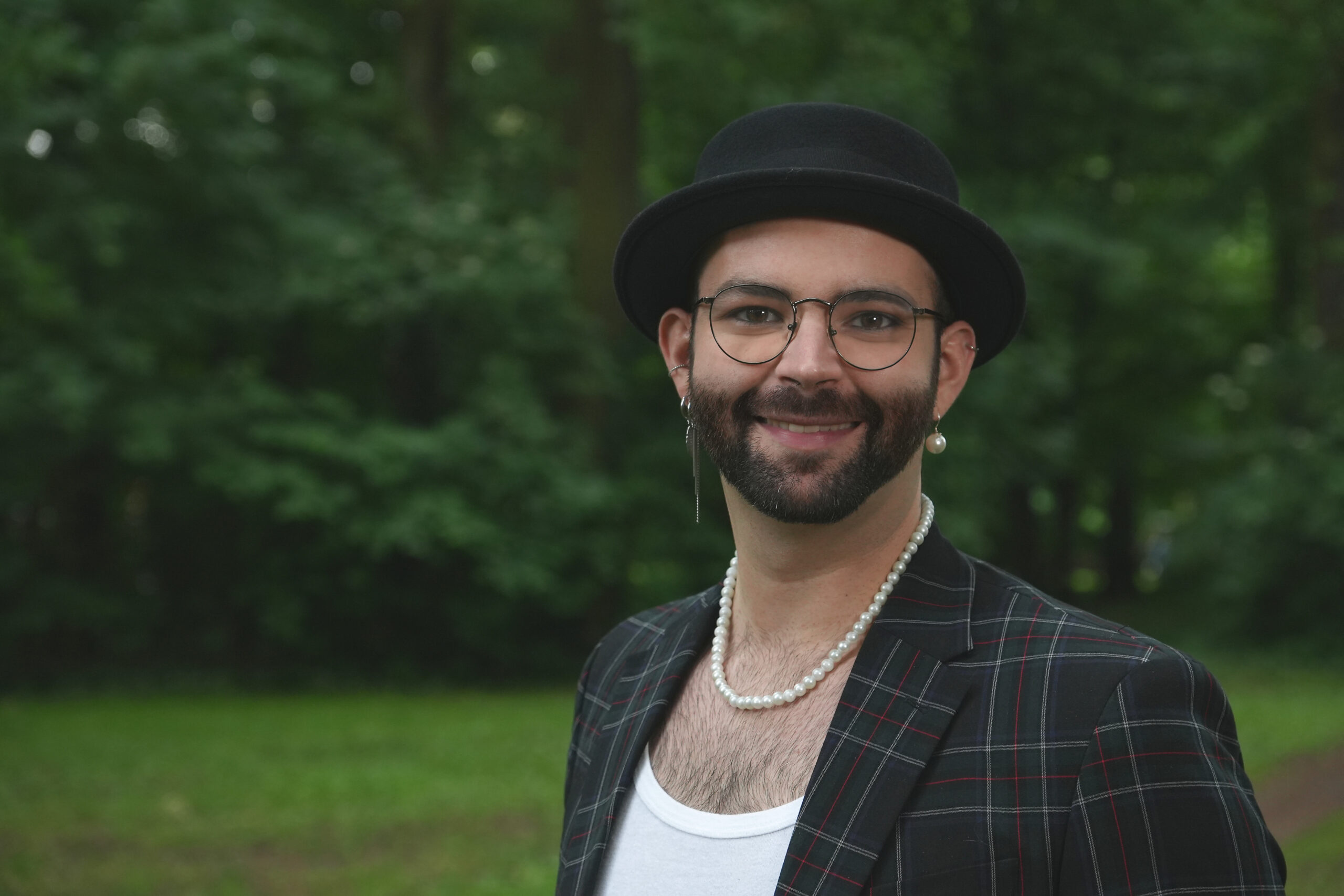 Ein dunkelhaariger, schlanker Mann zwischen 30 und 40 Jahren, mit Bart, schwarzer Brille, schwarzem Hut, Perlenkette, auffälligen Ohrringen und einen dunklen, karierten Jackett, steht auf einer grünen Wiese, man sieht den Oberkörper, im Hintergrund eine grüne Hecke.