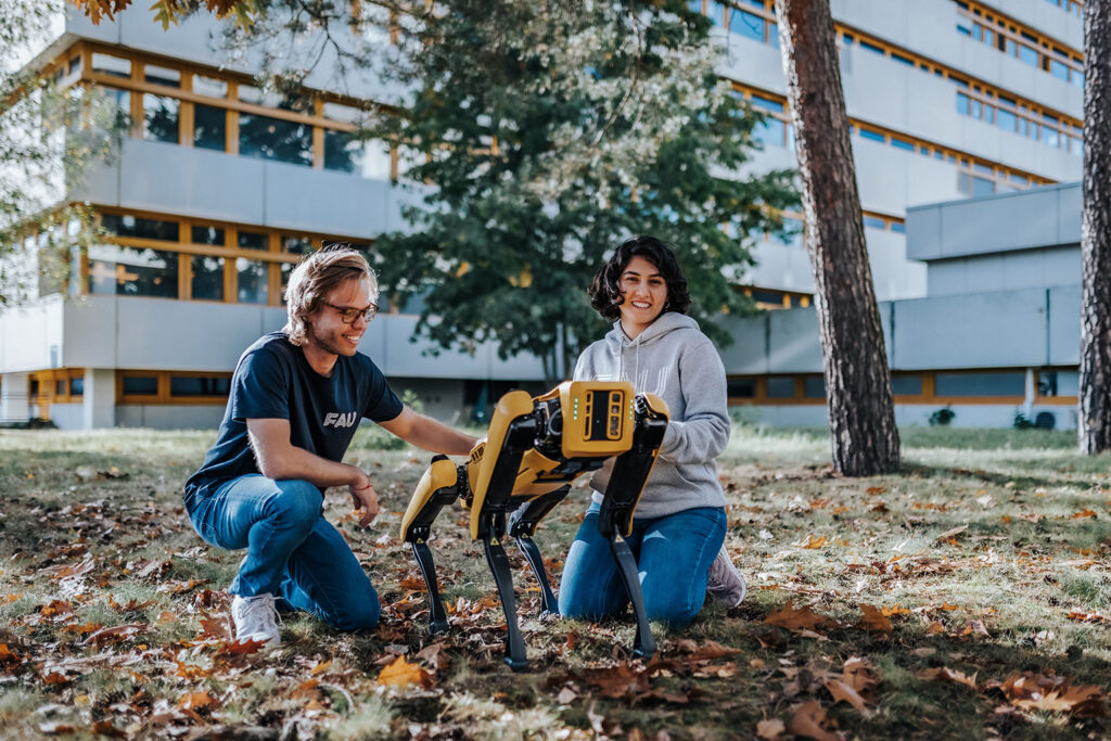 Zwei Personen sitzen mit einem vierbeinigen Roboter auf einer Campus-Wiese der FAU.