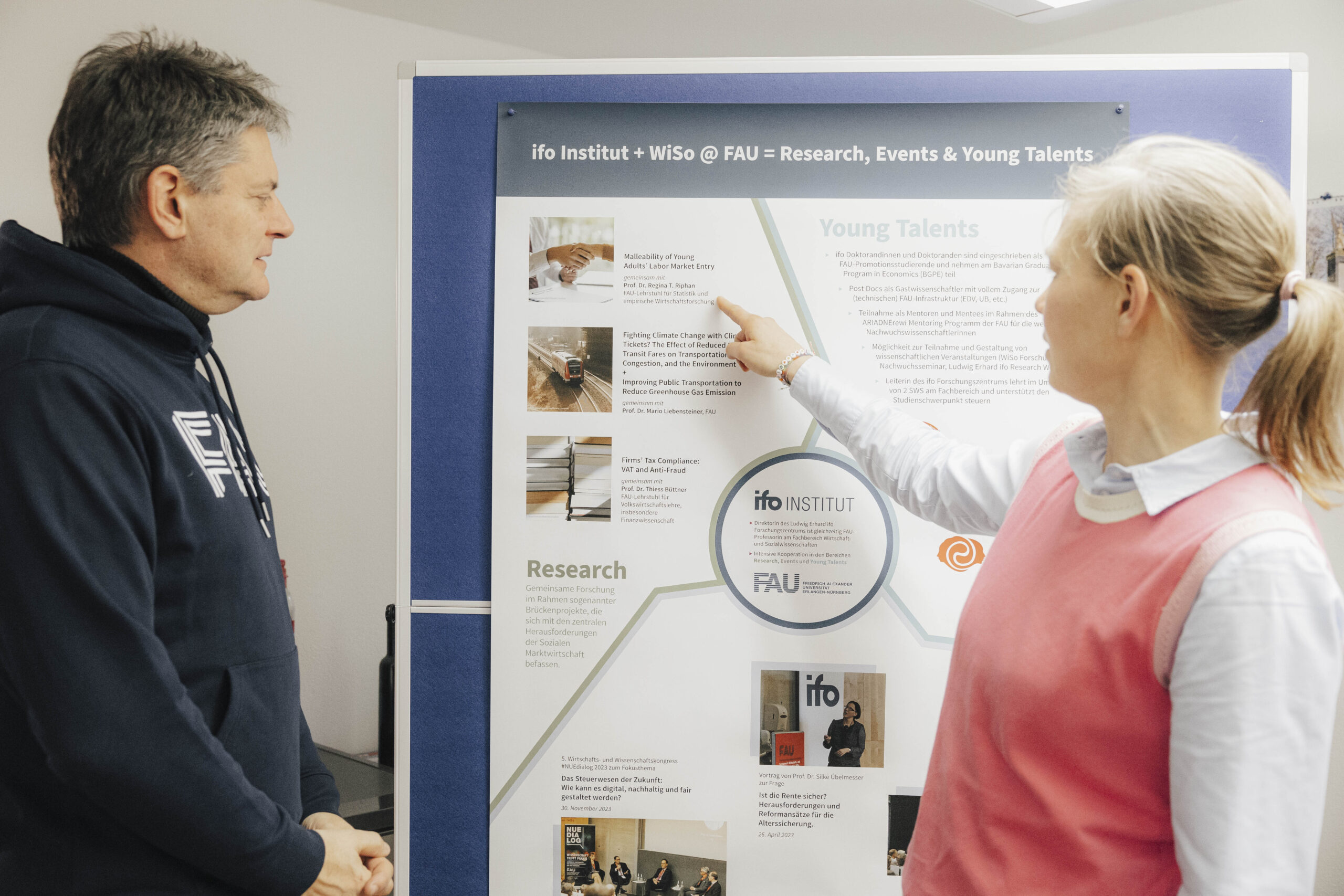 Der Unipräsident Hornegger und Professorin Necker stehen vor einer Pinnwand, Prof. Necker zeigt auf die Pinnwand, während sie etwas erklärt.