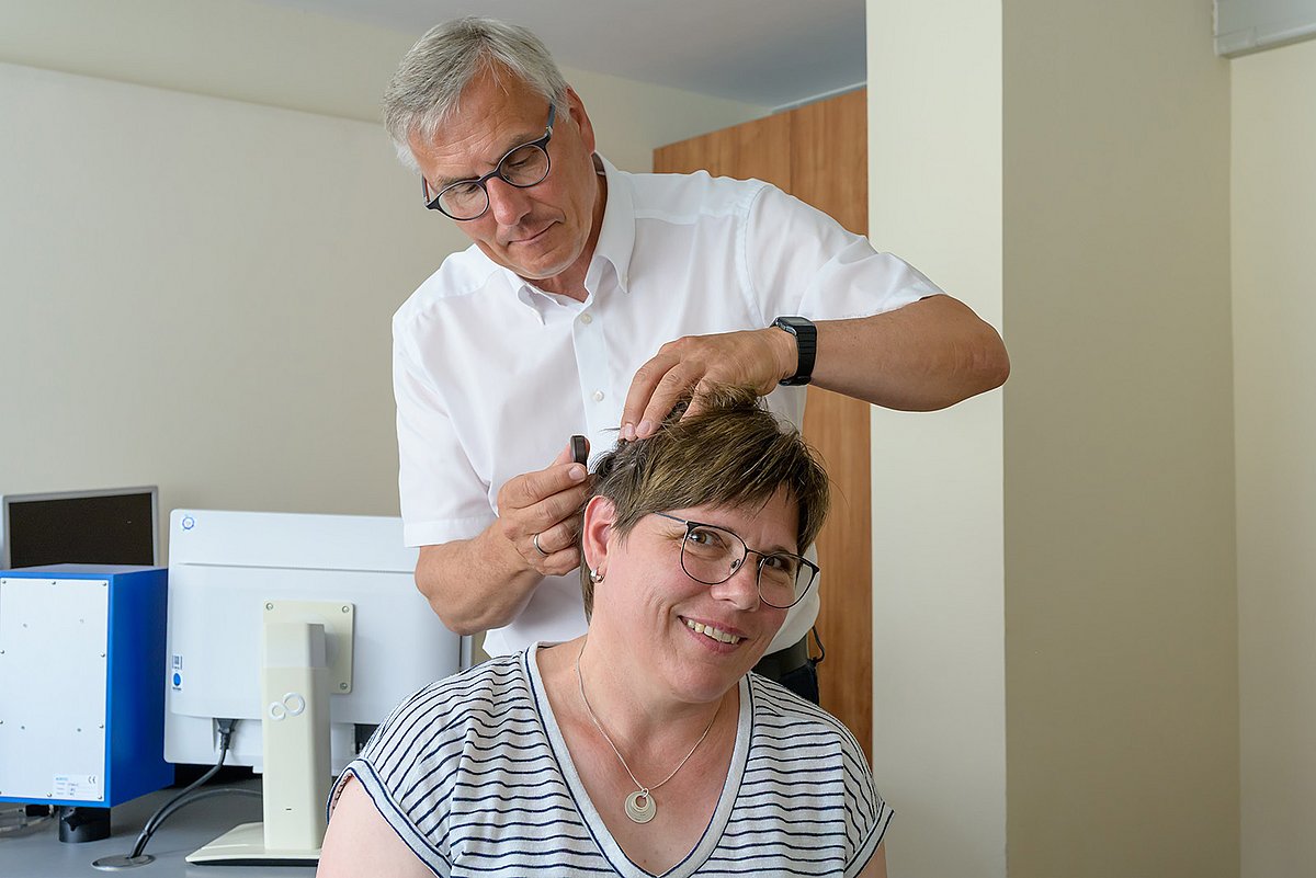 Prof. Hoppe im weißen Ärzteanzug setzt das Implantat ein. Die Patientin schaut freudig lachend in die Kamera.