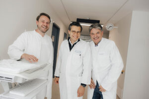 Prof. Dr. med. Ricardo Grieshaber-Bouyer und der Universitätspräsident Hornegger posieren zusammen lächelnd für ein Foto. Sie stehen in einem Krankenhausgang und tragen weiße Arztkittel, neben ihnen steht ein weiterer Mitarbeiter des Krankenhauses.