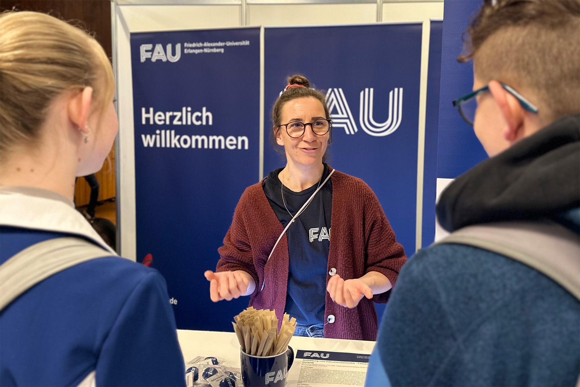 Eine Frau an einem Infostand der FAU berät zwei Menschen