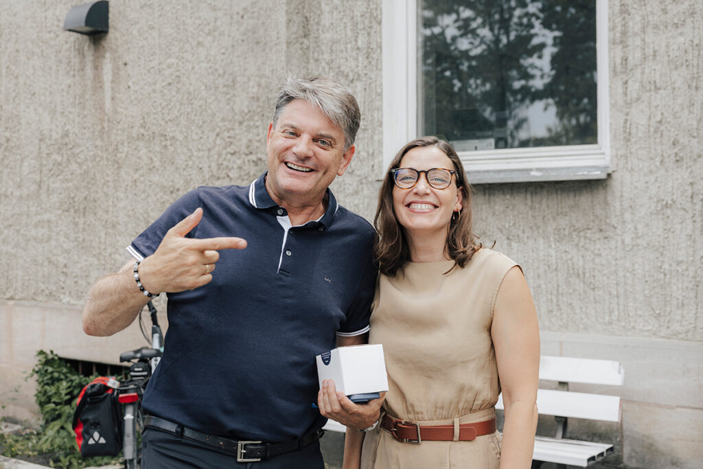 Präsident Hornegger und Prof. Kaifie-Pechmann stehen zusammen vor einem beigen Haus und lächeln in die Kamera. Hornegger hält in seiner rechten Hand eine kleine Verpackung, mit der linken Hand zeigt er auf Kaifie-Pechmann.