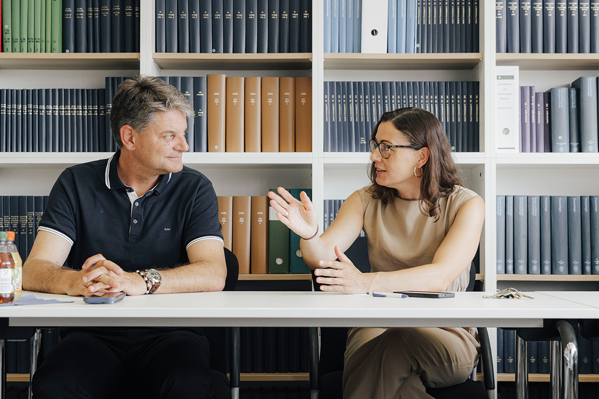 Hornegger und Kaifie-Pechmann sitzen nebeneinander an einem länglichen Tisch, im Hinterregal ist eine Bücherwand. Kaifie-Pechmann erklärt Hornegger etwas.