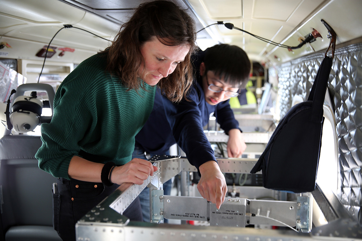 Ein Forscher deutet auf eine Vorrichtung in einem Flugzeug, im Vordergrund einen Wissenschaftlerin.