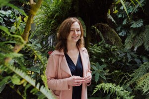 Prof. Jansen, eine Frau mit braunen haaren, trägt eine pinke lederjacke und steht im Botanischen Garten vor Bäumen und sträuchern.