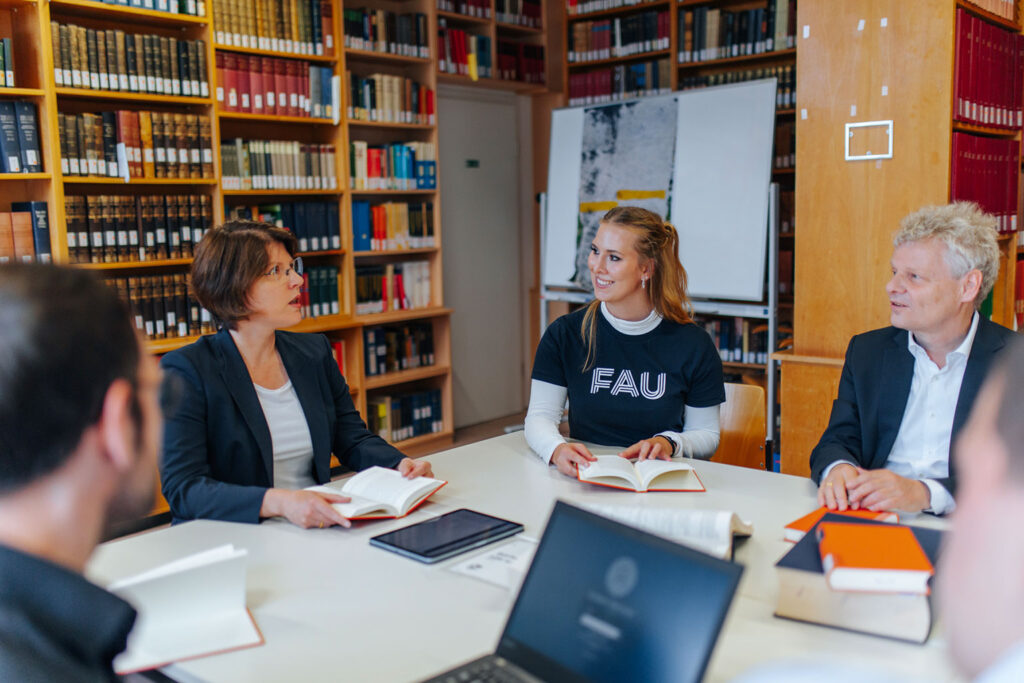 Mehrere Personen sitzen mit Büchern und Laptops in einer Bibliothek zusammen.